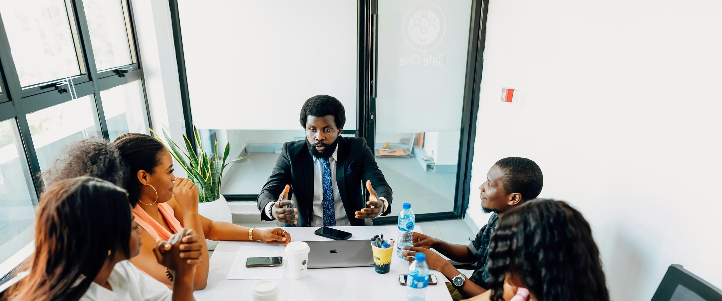 A team seated around a meeting table in a bright glass-walled office, listening to a colleague at the head of the table.