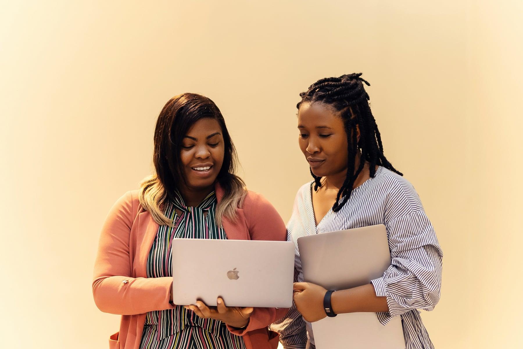 Two colleagues standing together, looking at an open laptop one of them is holding.